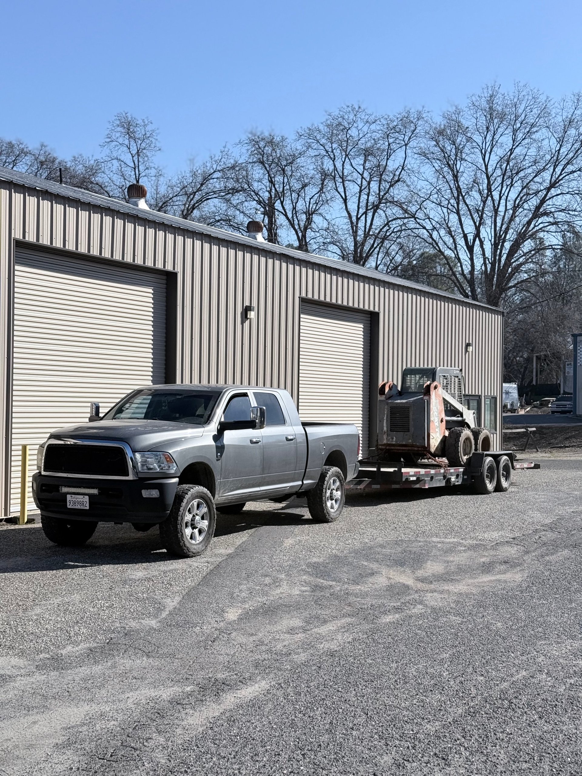 Silver pickup truck parked outside industrial metal warehouse with two white garage doors and bare trees in background